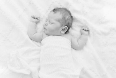 Black and white portrait of a newborn baby swaddled in a blanket atop a bedsheet with his hands and arms curled up