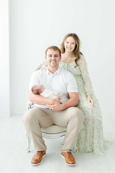 Father sitting in a chair holding newborn baby while mother leans behind them smiling during newborn photography session