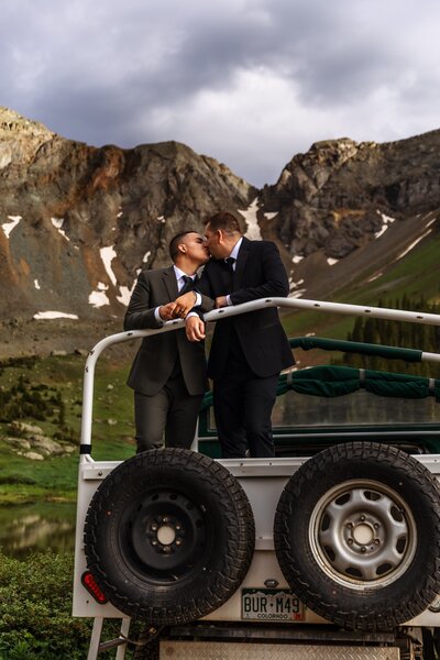 Shannon Lee, an adventure elopement photographer, captures a couple sharing a kiss on a rugged vehicle surrounded by a breathtaking mountain landscape.