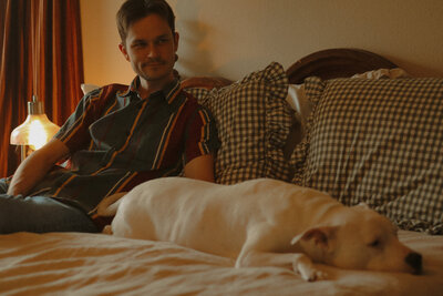 A man is sitting on a bed with checkered pillows and a lamp that is on in the background. His dog is laying on the bed next to him. Both are not looking at the camera.