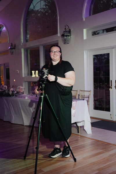 mara shields standing in front of the head table, with camera on a tripod, in a black dress and sneakers