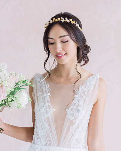 Kohnur Ethereal bride with natural bronzed makeup and long loose waves styled with a gold leaf and pearl hair vine, posing on a coastal cliffside.