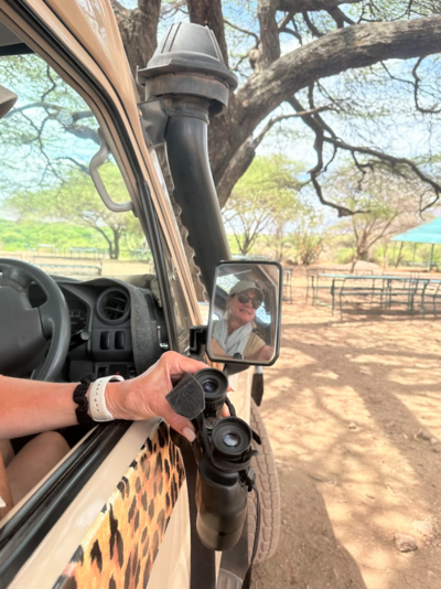 The reflection of a woman wearing sunglasses, a cap, and a scarf, is captured in the side mirror of a safari vehicle. The background reveals a dry, tree-filled safari landscape with picnic tables in the distance.