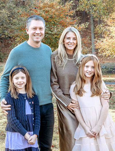 Stamford CT photographer creates stunning autumn family portrait with parents and two daughters wearing coordinated neutral tones against colorful fall foliage