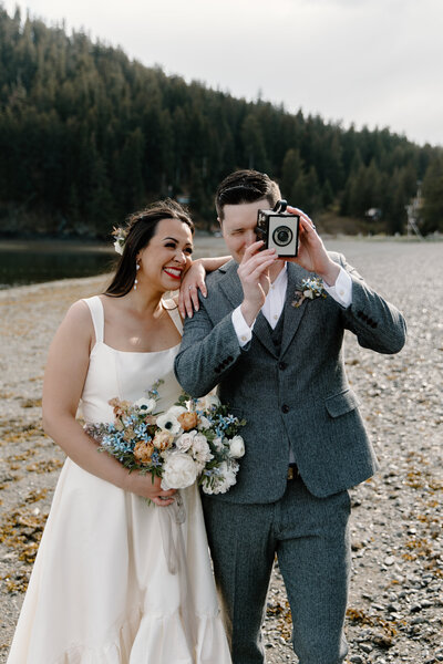 Groom holds film camera as bride smiles on Seldovia Alaska beach.
