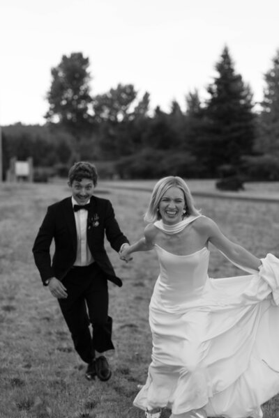 Bride and groom running hand in hand through grassy field in playful black and white portrait