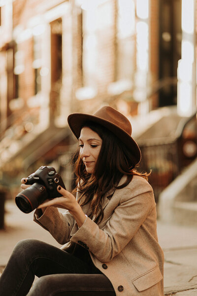 Woman sitting looking at her camera where a blazer and a hat on a New York City street.