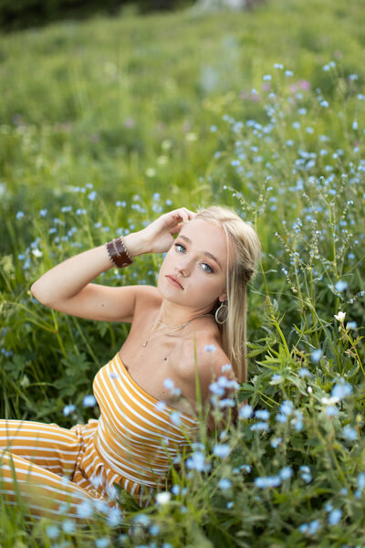 Young lady runs fingers through her hair as she directly looks forward while sitting in a field of wildflowers.