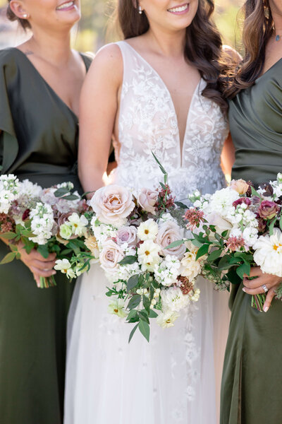 Three women in elegant dresses, with the central woman wearing a white floral gown and holding a bouquet of white and blush roses. The women on either side wear dark green dresses and hold similar bouquets. All are smiling, celebrating outdoors.