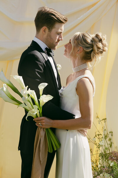 person in white dress and strapy heels standing against a white wall holding a white bouquet