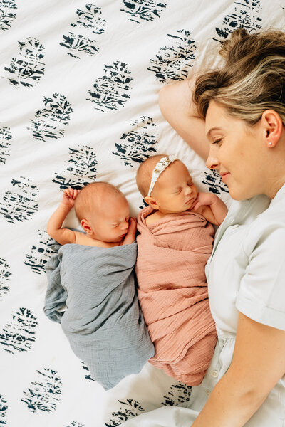 A mother lying on a bed with her twin newborns, both swaddled in soft blankets.