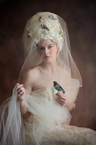 A girl wearing a marie antoinette wig  studded with birds is veiled with tulle while holding onto a bird