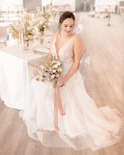 A woman sitting down in a white wedding dress holding a white floral bouquet.