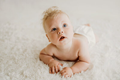 six month old baby boy lays on his belly on a white rug looking out the window while wearing a white diaper cover for his six month old Denver Mini milestone session