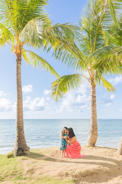 Photo of mother and daughter during an Oahu family photo session standing next to the water with palm trees framing the image.