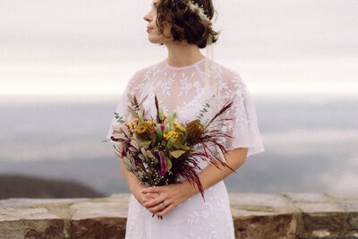 Bride holding a wildflower bouquet and gazing into the distance during her Virginia mountaintop elopement, captured in soft, editorial light with a film-inspired look.


