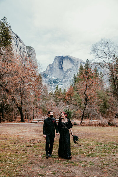 A couple dressed in dark wedding attire walks hand-in-hand through a grassy meadow in Yosemite, smiling warmly at each other with the iconic Half Dome towering majestically behind them