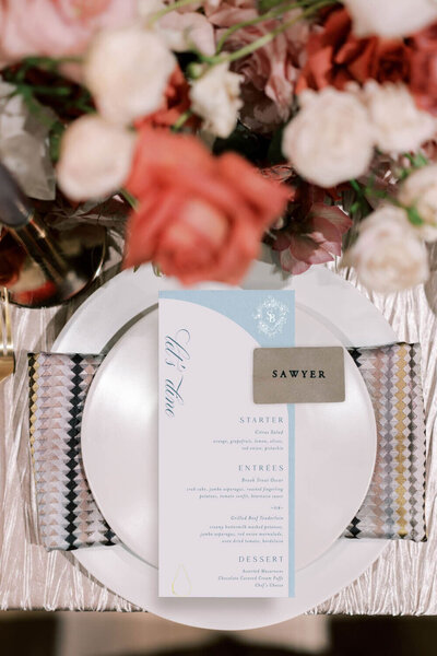 Table setting with a modern wedding menu printed on curved stationery, geometric napkin, and custom name card, surrounded by warm-toned floral arrangements.