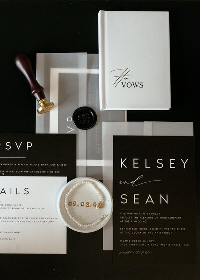 A stylish wedding invitation suite with black and white stationery, gold wax seal, and vow booklet, paired with a pearl bracelet and elegant wedding date details on a ceramic dish.