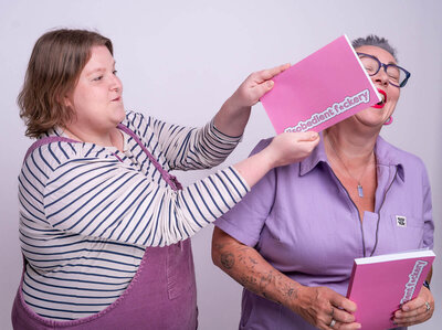 In a studio, two people play around. Lucy on the left, wearing a striped top with mauve dungarees, motions a bright pink notebook labelled “disobedient f*ckery” toward Pippa's face -  a cheeky grin on their face. Pippa on the right in lilac collared dungarees, blue rimmed glasses, tattooed forearms, laughs while holding a second same pink notebook.
