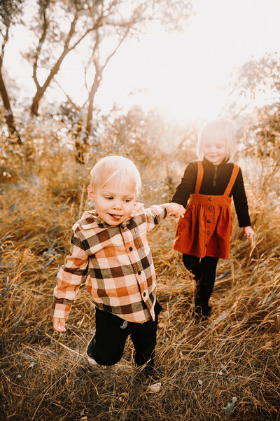 Little brother holds tight to big sisters hand as they walk through the prairie grass at Sweet Briar Lake during their Fall Mini Session with Nikii Pix Photography of Bismarck