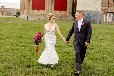 A bride and groom hold hands, captured by a talented Minnesota photographer, as they stroll through a grassy area. The bride's white gown and vibrant bouquet of pink and purple flowers complement the groom's dark suit with a floral boutonniere. A rustic building adds charm to this beautiful wedding photography moment.