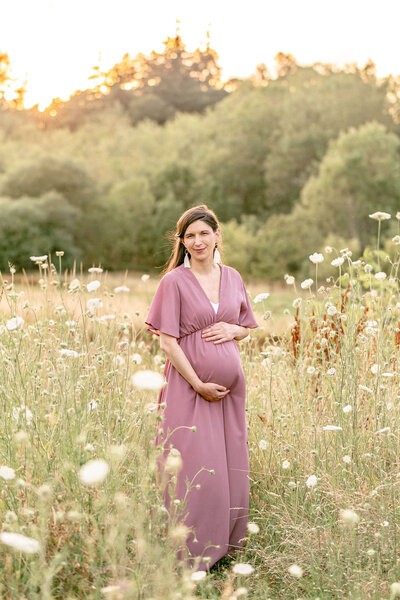 Woman in mauve dress holding pregnant belly while standing in a field of wildflowers.