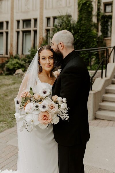 In this enchanting outdoor moment, a smiling bride showcases her modern bouquet in soft earth tones while her groom kisses her forehead affectionately. Their joyful connection captures the essence of love, perfect for couples in Ottawa looking for personalized wedding floral arrangements and heartfelt moments.