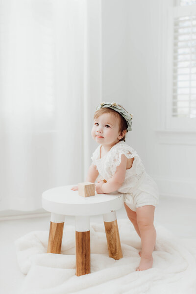 Baby girl in white lace outfit stands at a stool during baby photography session in Raleigh NC