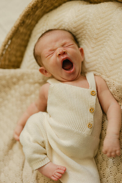 A newborn baby yawns while sleeping in a woven basket while wearing a knit onesie in the studio of a Mississauga Newborn Photographer