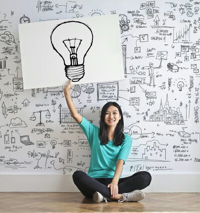 Attractive woman holding a poster with a lightbulb and many visuals around strategy on the wall behind her