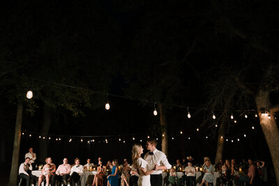 Newlyweds dance under the moonlit sky with cafe lights surrounded by their family and friends.