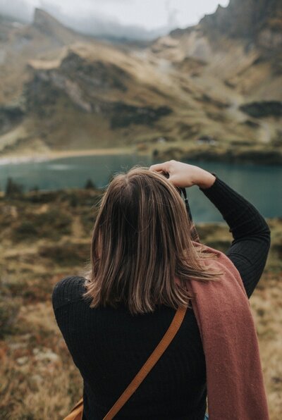 photographer is holding a camera with her back facing the camera. she is taking a photo of an alpine lake and mountains. It is fall and moody. The grass is brown and the lake is blue.
