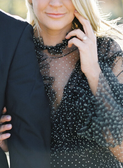 Luxury engagement session. He is in a navy dinner jacket, but only his arm and shoulder are  in the photograph. She has blonde hair that is straight and down and blowing gently with the wind. She has on a black collared dress with long sleeves and small white polka dots. The dress has a sheer V from the ruffled collar to the bottom of the chest. Her sleeves are sheer as well with ruffles at the wrists.  Her right hand is linked through his arm and her left hand is up by her face showing off the ring. Photographed by wedding photographers in Charleston Amy Mulder Photography