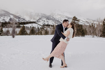 Wyoming elopement with groom holding his bride around the waist and kissing her as she pops her foot up in the air in a snow filled field with the mountains covered in snow in the distance  photographed by photographers Jackson Hole