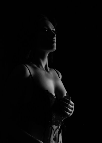 Black and white photo showcasing a woman in lingerie, lit from the side, creating dramatic shadows that highlight her silhouette. She is looking upwards and holding her torso, emphasizing the contours of her body against the dark background.