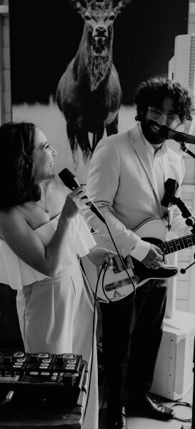 Black-and-white image of a female vocalist and male guitarist performing as a duo indoors, with a stag mural in the background.
