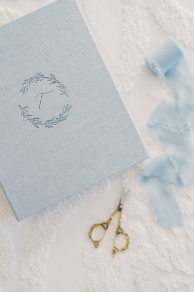 Light blue linen box lays next to blue ribbon and gold scissors.