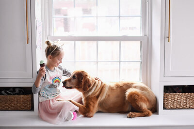 Little girl with a tiara and ice cream in her hands sits at the window with a golden retriever begging for a lick of the ice cream.