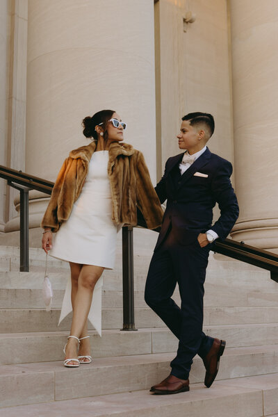A wedding couple standing on steps leaning against a railing looking at each other. The bride is wearing a above knee white dress with a brown fur coat and white sunglasses, and the groom is wearing a black tuxedo with brown shoes.