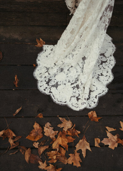 Lace from a white wedding dress laying across a dark deck next to a yellow pile of leaves. 