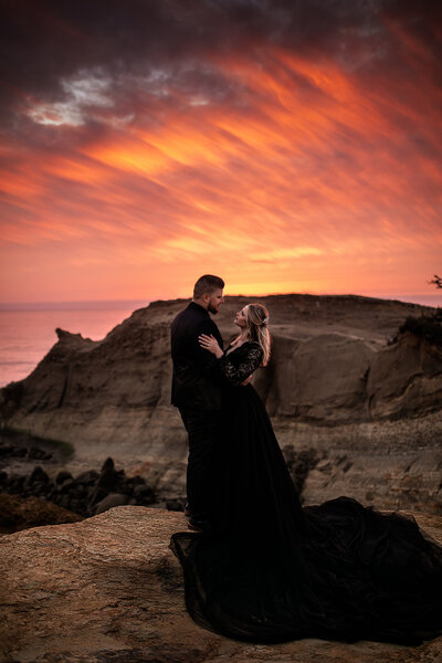 A couple in black attire stands on a cliff at sunset with vibrant orange skies, captured by an Oregon elopement photographer.