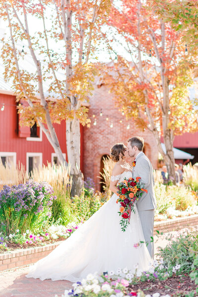 A bride and groom kiss on a brick pathway surrounded by flowers in front of a red barn wedding venue.