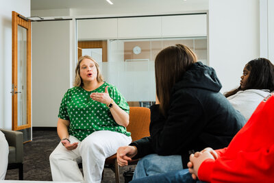Mya Anderson in a green polka dot shirt speaks to a group of students seated in a modern, bright room during The Summer Jumpstart Program, helping students get college-ready this summer.