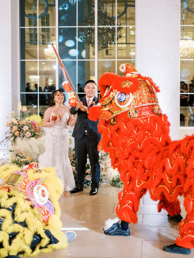 A Chinese red dragon dances across the floor in front of a chinese bride and groom who hold wine and a lantern on a stick.