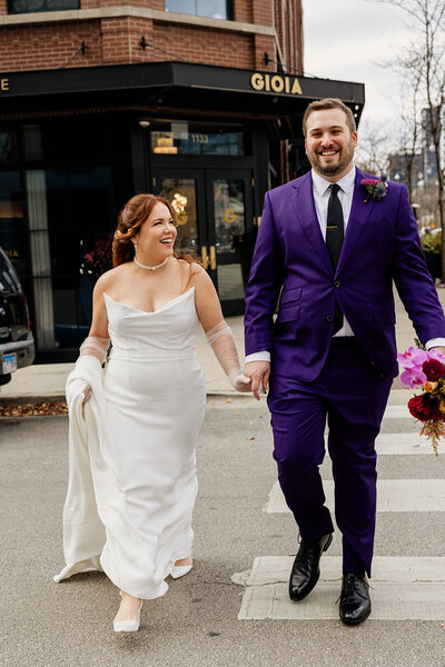 Red-headed bride in timeless, strapless gown smiles at groom wearing a purple suit while holding hands and crossing the street.