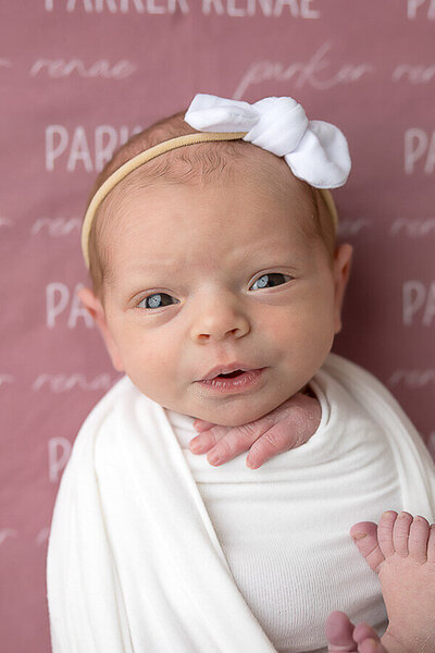 Baby girl with white bow on a pink blanket looks into camera.