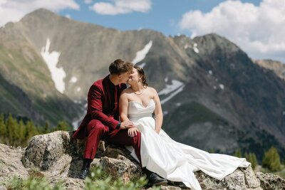 Bride and groom kiss while sitting on rock in front of towering mountains in Colorado elopement day.