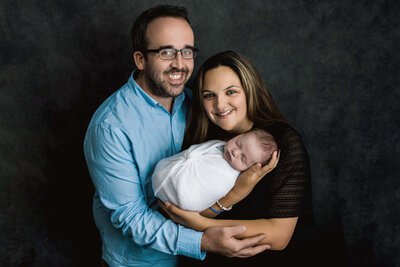 Joyful new parents smiling while holding their sleeping newborn baby wrapped in white during professional studio portrait session on dark backdrop