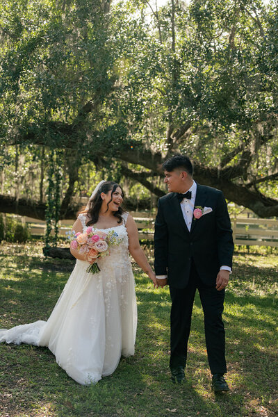 Bride and groom holding hands and smiling under a canopy of oak trees at Uliana Estate in Lakeland, Florida during summer